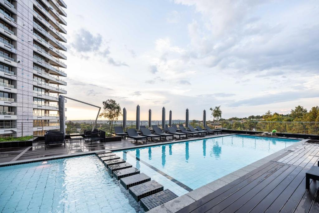 Piscina moderna na cobertura do Sandton Hotel com espreguiçadeiras, móveis pretos à beira da piscina e vista da cidade sob um céu parcialmente nublado. A foto faz parte do post de onde ficar em Joanesburgo.