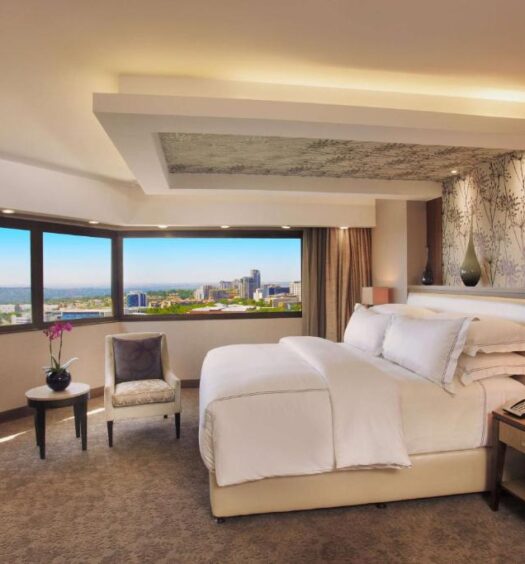 Quarto do hotel Sandton Sun and Towers com uma cama grande, TV de tela plana, área de estar e vista da cidade através de janelas amplas. A foto é capa do post de onde ficar em Joanesburgo.