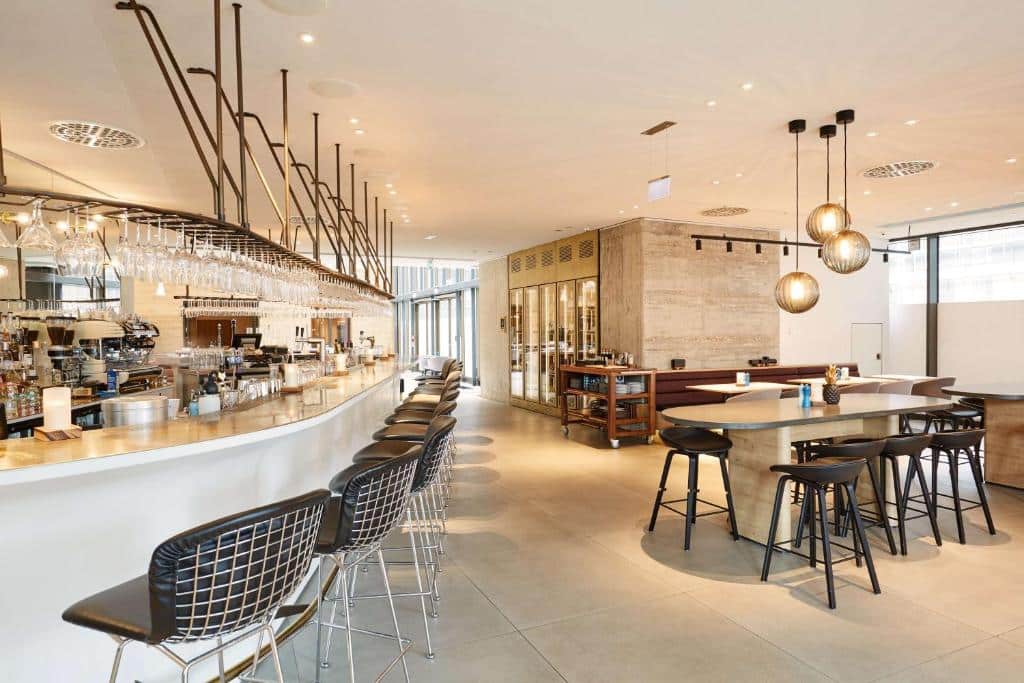 Foto do Scandic Frankfurt Museumsufer. Bar moderno e área de jantar com banquetas altas, mesas redondas, luzes penduradas e um balcão com copos e garrafas.