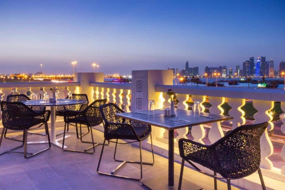 Terraço externo de um restaurante no Souq Waqif Boutique Hotels com quatro mesas e cadeiras modernas com vista para o horizonte da cidade ao anoitecer.