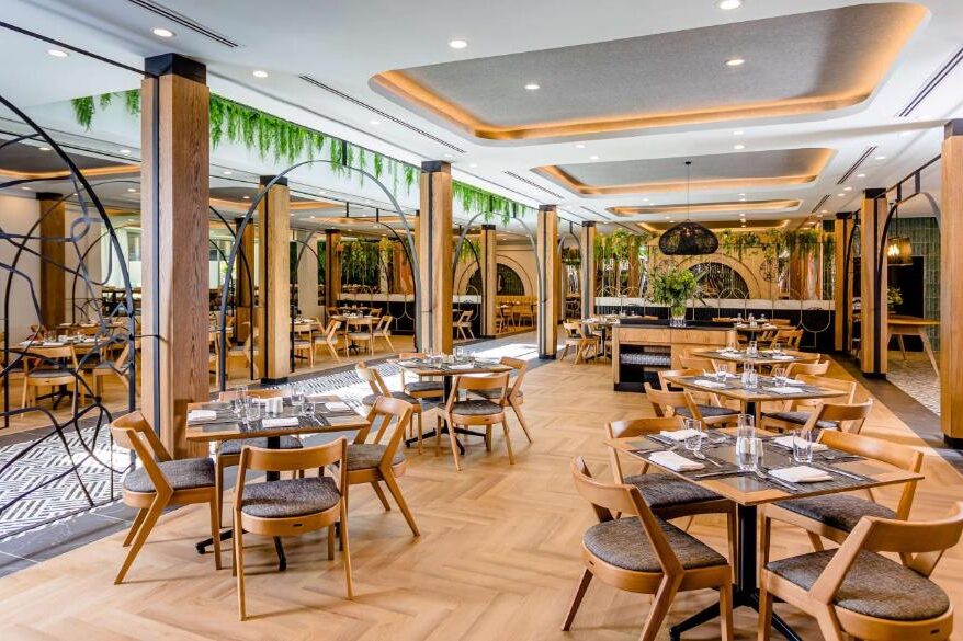 Interior de restaurante do Southern Sun Rosebank com mesas e cadeiras de madeira, vegetação, janelas grandes e iluminação decorativa. A foto é capa do post de onde ficar em Joanesburgo.