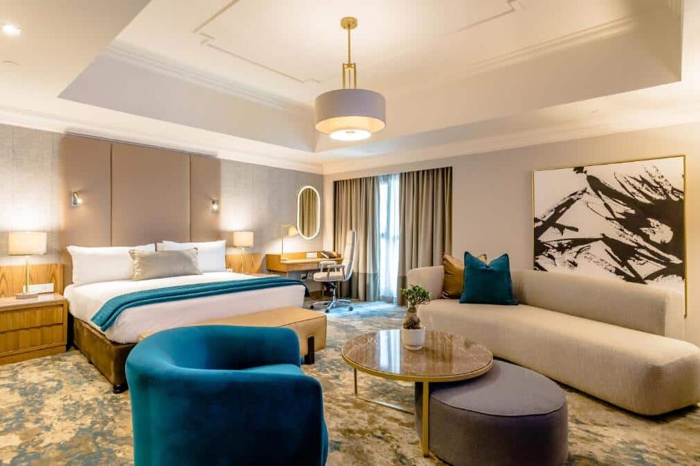 Quarto do Southern Sun Sandton com uma cama king size, sofá, poltrona azul, escrivaninha, arte abstrata e janela grande com cortinas. A foto faz parte do post de onde ficar em Joanesburgo.