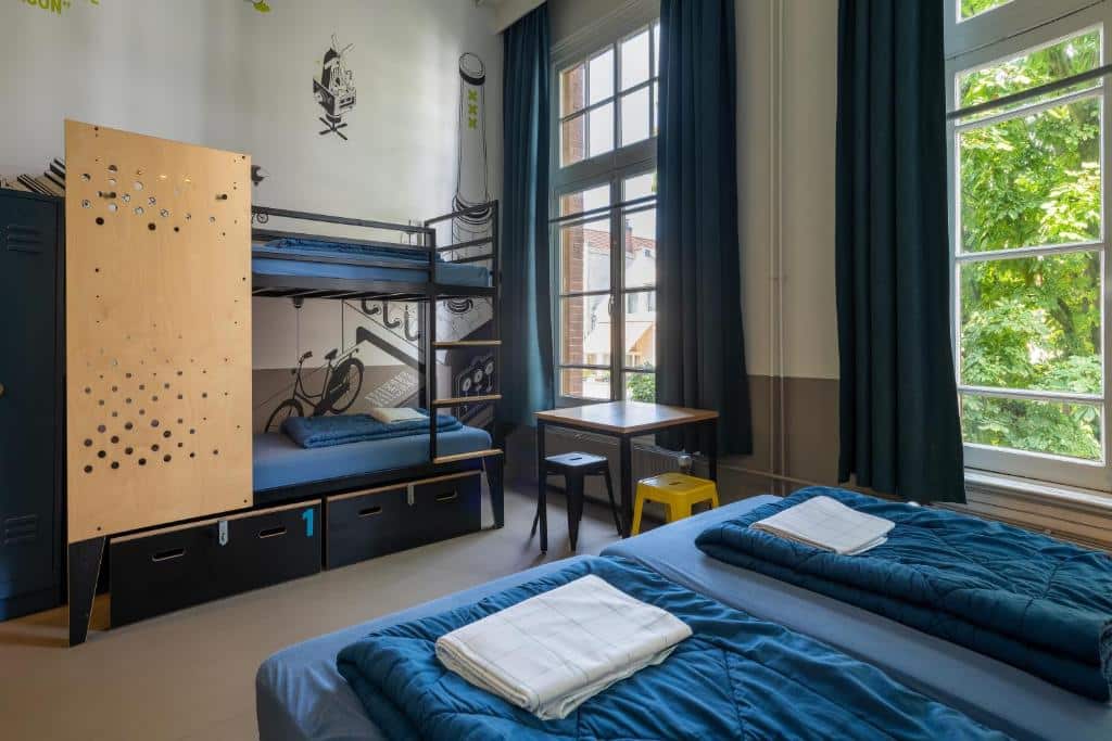 Foto do Stayokay Hostel que ilustra onde ficar em Amsterdam. Um quarto de albergue com um beliche, duas camas de solteiro, armários, uma escrivaninha, janelas grandes e toalhas dobradas sobre as camas.