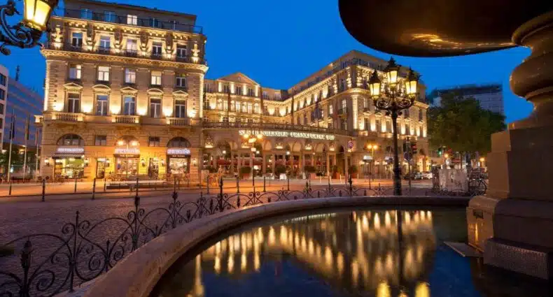 Edifício histórico com janelas iluminadas ao anoitecer, visto de uma fonte com reflexos e lâmpadas de rua ornamentadas.