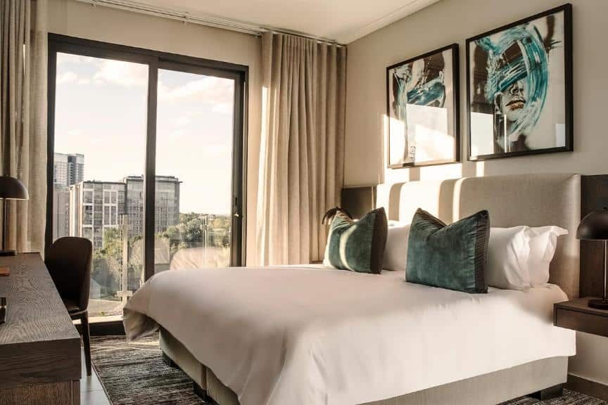 Quarto do The Catalyst Apartment Hotel com uma cama de casal, janela grande, escrivaninha, duas luminárias e obras de arte abstratas acima da cama. A foto faz parte do post de onde ficar em Joanesburgo.