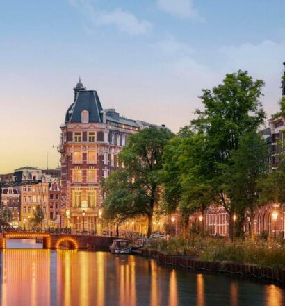 Um canal em uma cidade repleta de árvores e edifícios históricos, iluminado por postes de luz ao anoitecer.