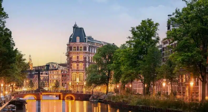 Um canal em uma cidade repleta de árvores e edifícios históricos, iluminado por postes de luz ao anoitecer.