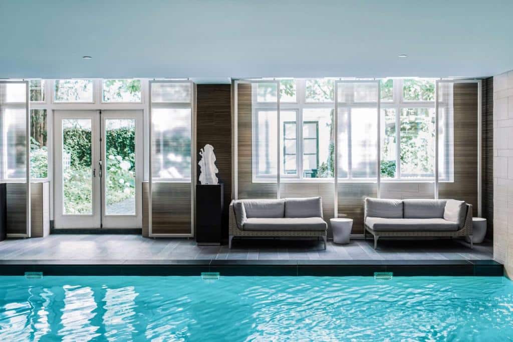 Foto do Waldorf Astoria Amsterdam. Área da piscina interna com água azul clara, dois sofás cinza, mesas laterais brancas e janelas grandes que permitem a entrada de luz natural.