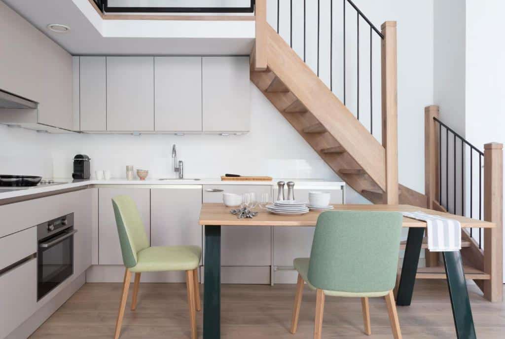 Cozinha moderna do apartamento Wittenberg by Cove, com armários de madeira clara, uma pequena mesa de jantar para quatro pessoas e uma escada de madeira com corrimão preto.