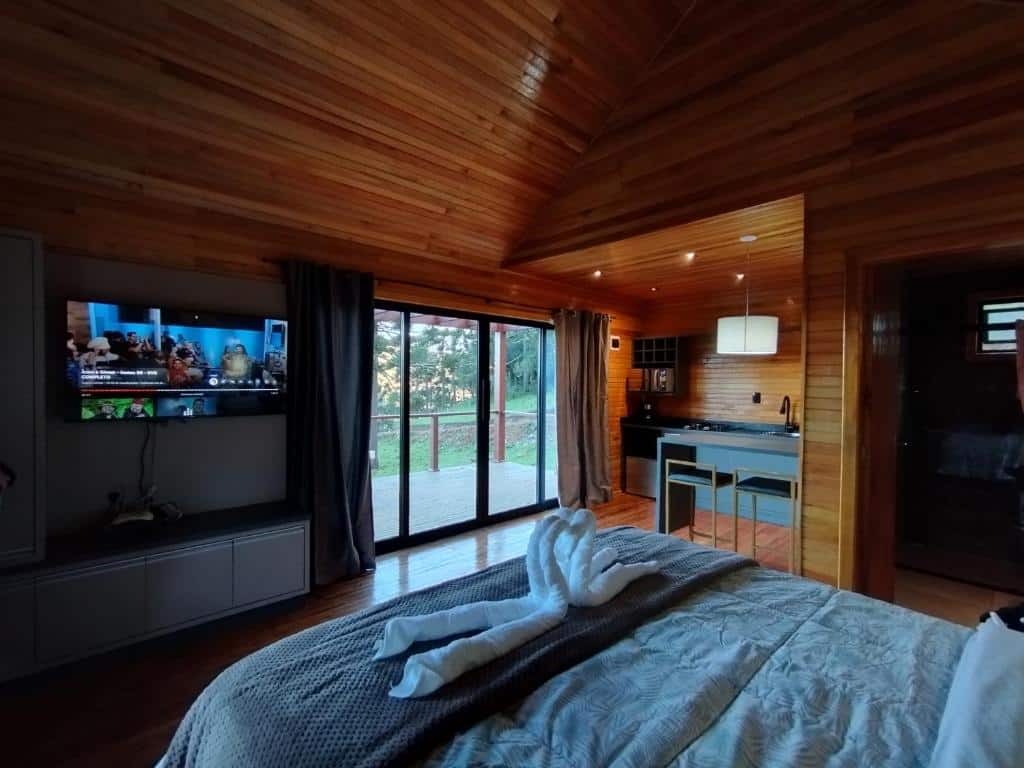 Um quarto em cabine de madeira da Monte Alegre Pousada, com TV, cama, toalha de cisne, portas de vidro deslizantes e uma pequena cozinha.