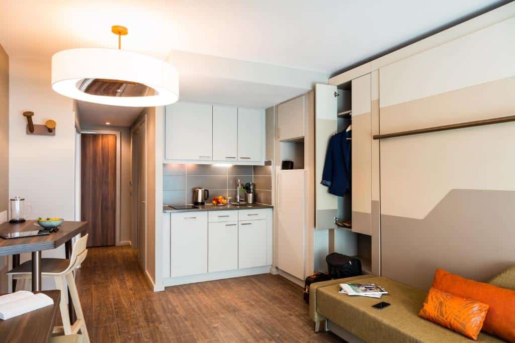 Aparthotel Adagio Edinburgh Royal Mile moderno com cozinha compacta, armário aberto, sofá e uma pequena área de jantar com detalhes em tons neutros e laranja.