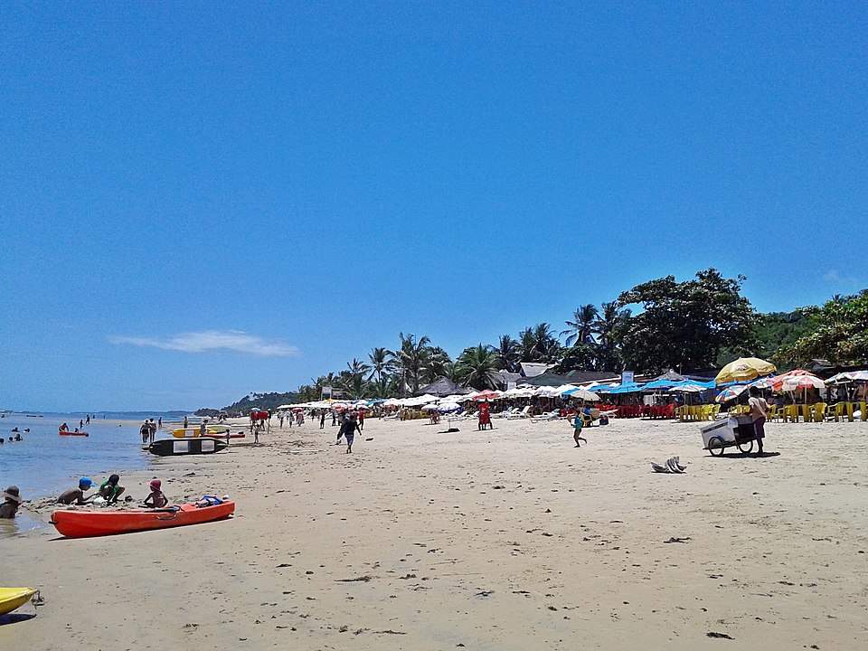 As pessoas relaxam em uma praia de areia com guarda-sóis, palmeiras e céu azul. Alguns nadam, outros caminham ou se sentam sob os guarda-sóis, na Praia do Mucugê, Arraial d'Ajuda.
