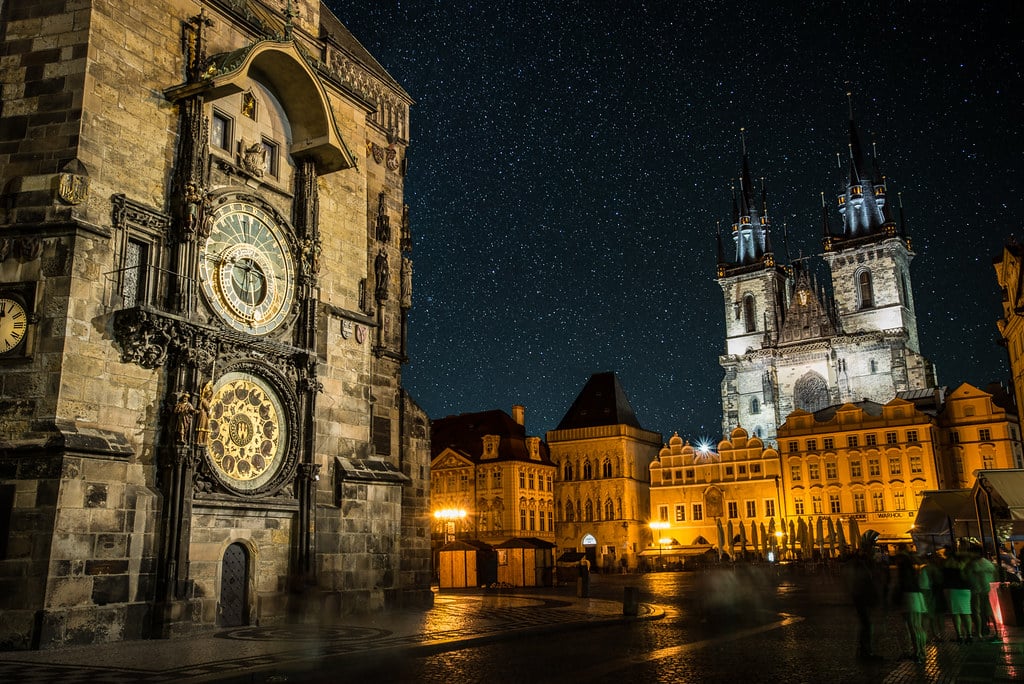 Foto noturna do centro histórico de Praga. A torre do relógio astronômico está à esquerda, e no centro da foto mais ao fundo há outros edifícios históricos iluminados por uma luz quente. O céu está bastante estrelado.