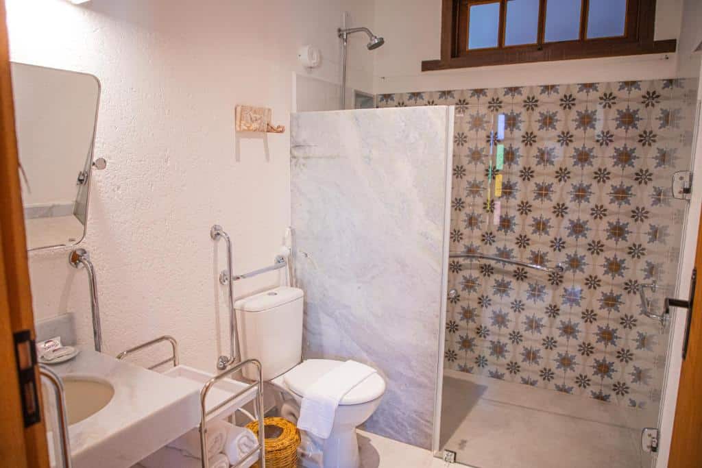 Banheiro acessível do Tororao Hotel Pousada com barras de apoio, box amplo, parede de azulejos estampados, vaso sanitário, pia e uma janela acima do chuveiro.