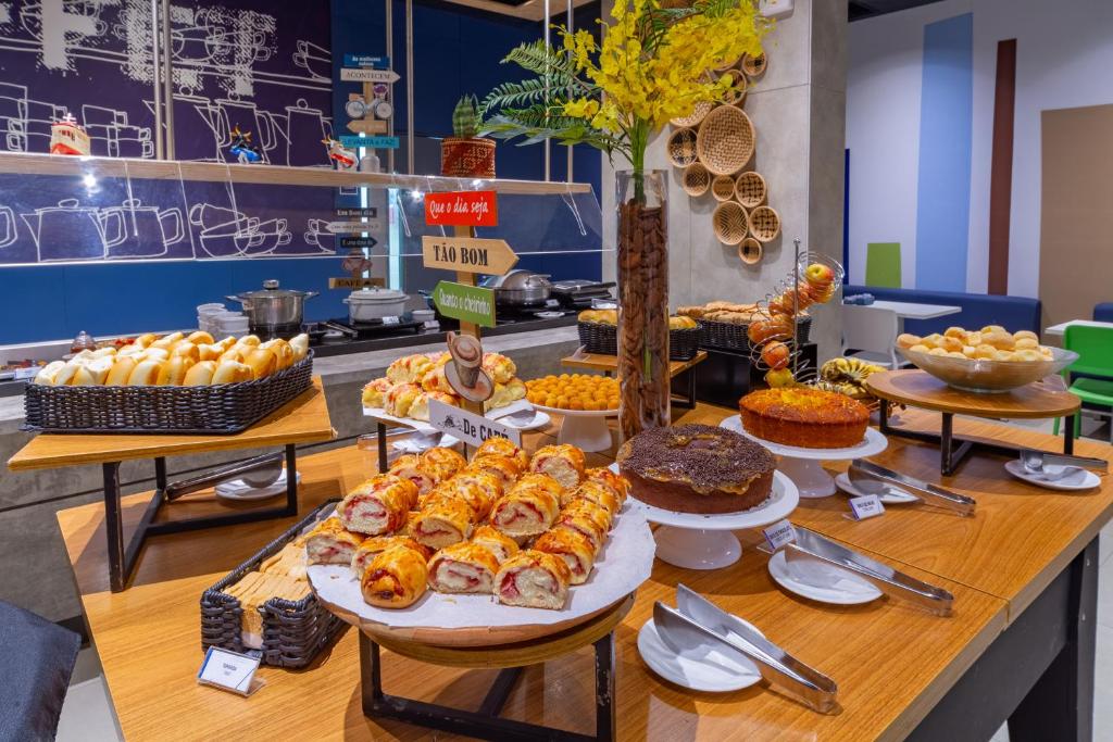 Uma mesa de bufê com pães, doces, bolos e tigelas variados, com utensílios de servir e flores amarelas decorativas, do ibis budget Manaus. Representa hotéis em Manaus.