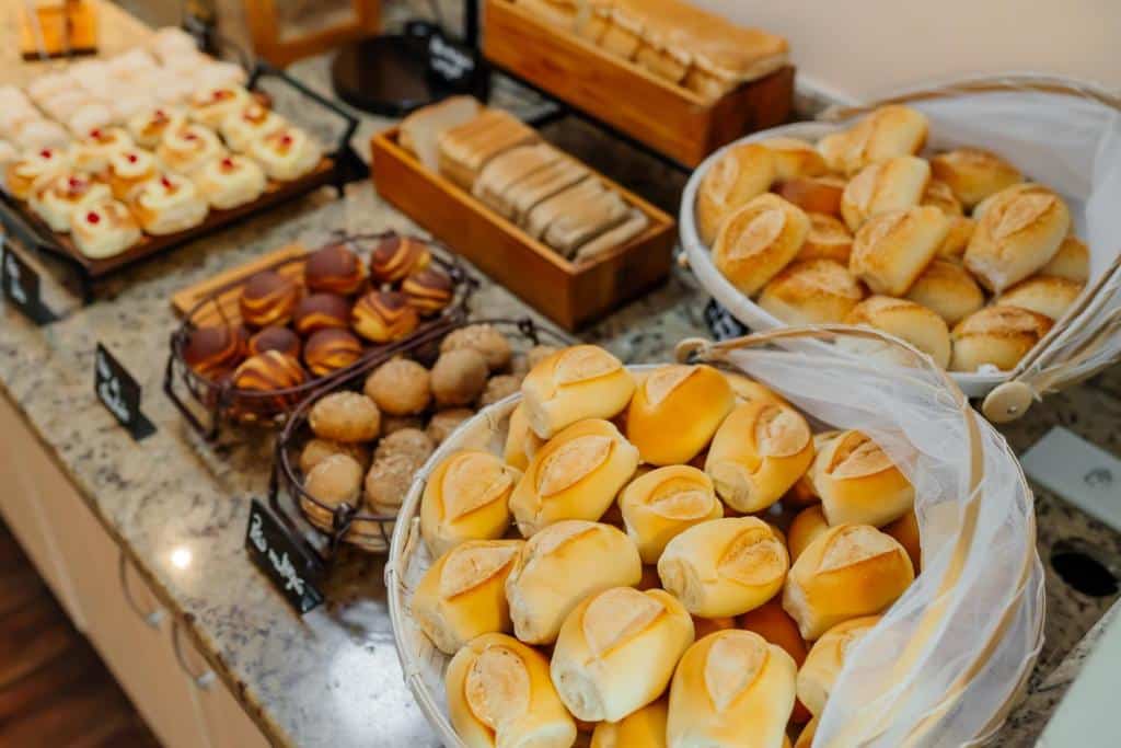 Pães e doces variados são exibidos em cestas e bandejas em um balcão de granito em uma padaria ou bufê, do ibis Styles Manaus. Representa hotéis em Manaus.