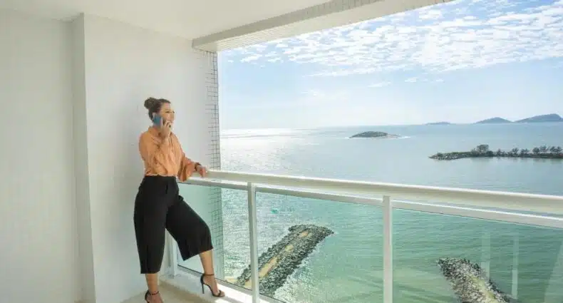 Mulher em pé em uma varanda à beira-mar, falando ao celular, com vista para a água e ilhas distantes, no Golden Tulip Macaé. Representa hotéis em Macaé.