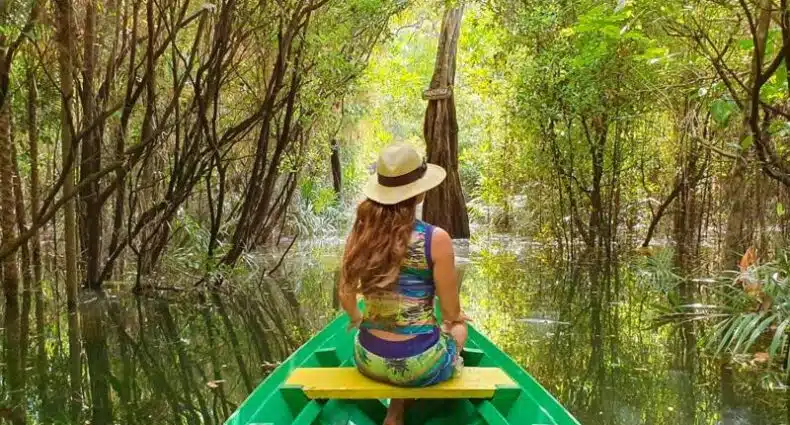 Uma mulher de chapéu senta-se à frente de um barco verde, viajando por uma floresta inundada com densa folhagem verde, em Hotel Terra Nativa. Representa hotéis em Alter do Chão.