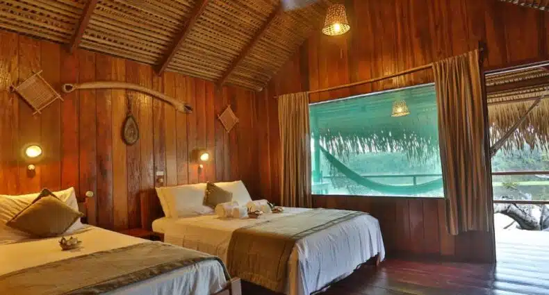 Duas camas de casal em um quarto rústico de madeira com uma grande janela, roupa de cama bege e teto de tecido, com vista para uma rede do lado de fora, no Juma Amazon Lodge. Representa hotéis em Manaus.