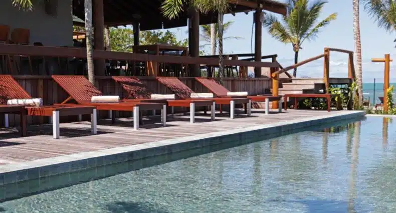 Vista da piscina com várias espreguiçadeiras vermelhas e toalhas enroladas em um deck de madeira, plantas e árvores tropicais ao fundo, em Santa Aldeia Hotel. Representa hotéis em Arraial d’Ajuda.