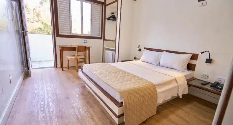 Um quarto do Sesc Contagem arrumado com uma cama de casal, escrivaninha e cadeira de madeira, ganchos de parede e uma janela que permite a entrada de luz natural. Representa hotéis em Contagem.