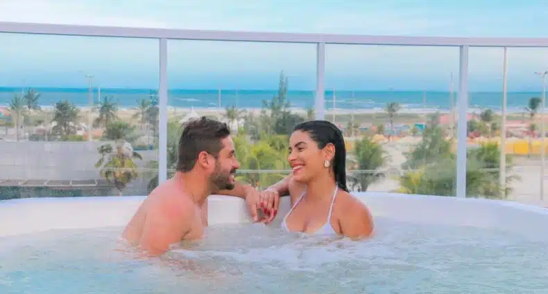 Um homem e uma mulher sentam-se e sorriem um para o outro em uma banheira de hidromassagem na cobertura, com uma praia e palmeiras ao fundo,em Vidam Hotel Aracaju.