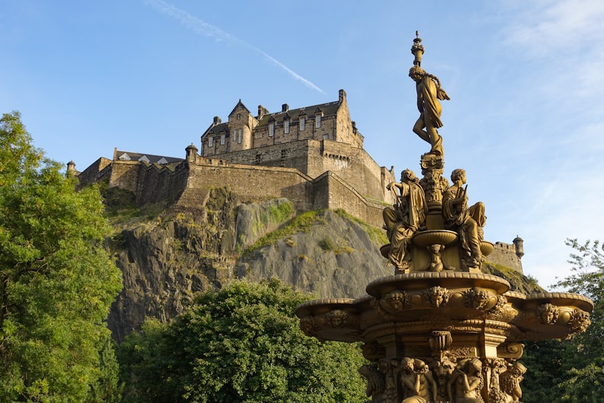 O Castelo de Edimburgo fica no topo de uma colina rochosa, com uma fonte decorativa em primeiro plano e árvores ao redor da cena.