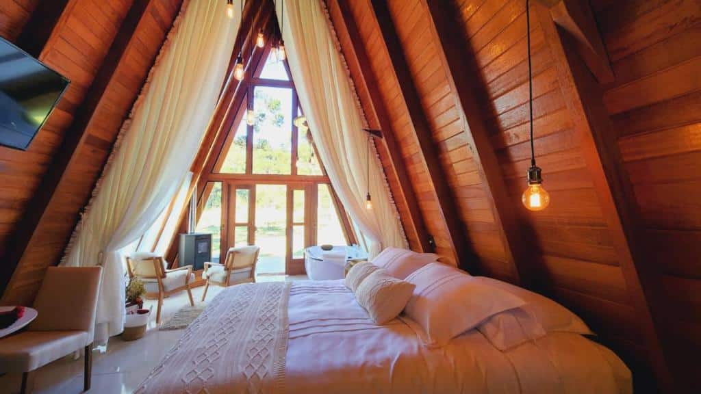 Interior do chale da Pousada Maçã Araucária, em São Joaquim, em forma de cabine (triângulo) com painéis de madeira, uma cama grande, uma TV, poltronas e luz do sol que entra pelas janelas altas.