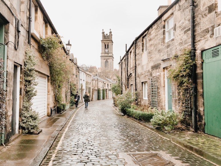 Uma rua estreita de paralelepípedos ladeada por edifícios de pedra, vegetação e uma torre de igreja visível ao fundo.