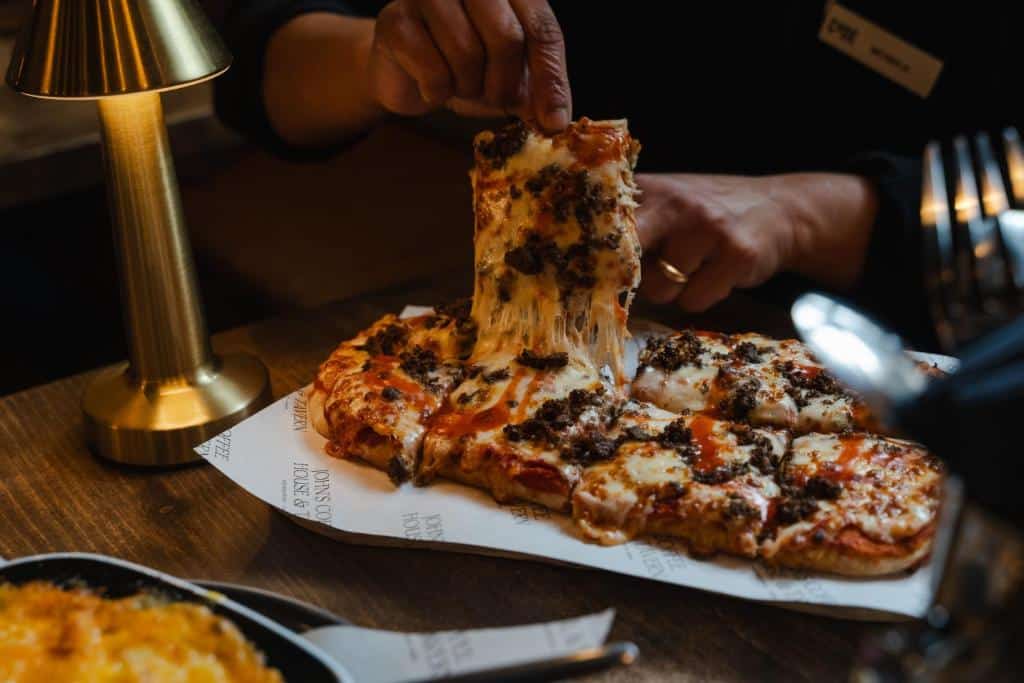 Uma pessoa levanta uma fatia de queijo de uma pizza com cobertura de carne em uma mesa de madeira em um ambiente pouco iluminado no Code Pod - The Court. Ilustra o post de onde ficar em Edimburgo.
