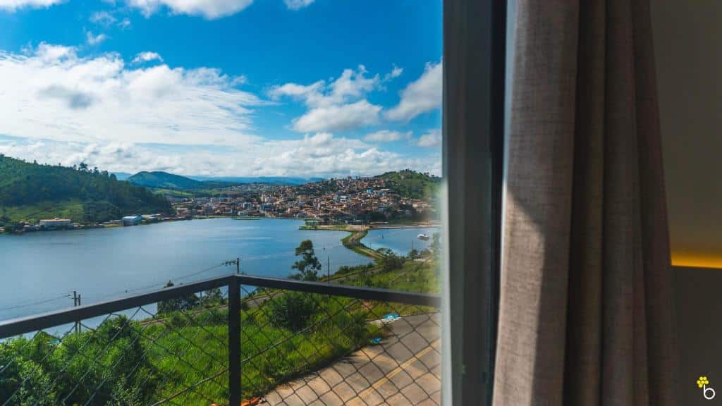 Vista de uma janela mostrando uma varanda, um lago, vegetação e uma cidade sob um céu azul parcialmente nublado, do NAIM Hotel Capitólio.