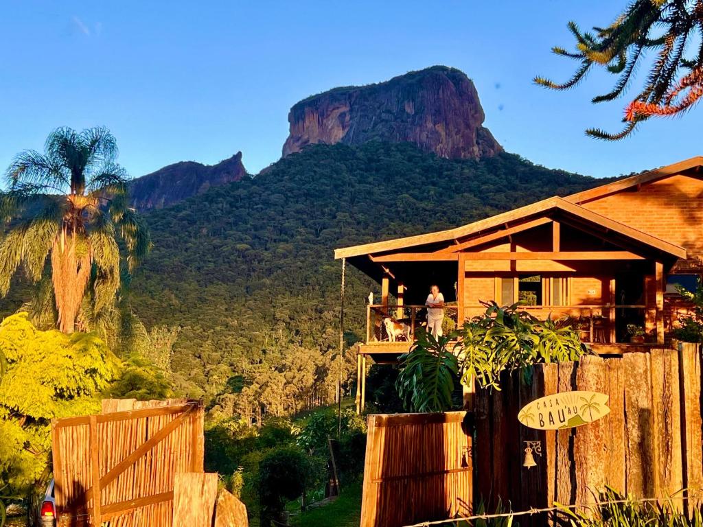 Cabana de madeira da Casa Baúau com varanda com vista para uma floresta verde exuberante e uma grande montanha rochosa sob um céu azul claro. Representa hotéis em São Bento do Sapucaí.