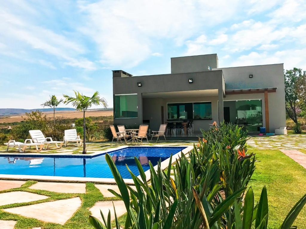 Casa moderna com pátio, piscina, cadeiras de gramado e plantas em um dia ensolarado com céu azul e colinas ao fundo, em Casa Lago de Furnas.