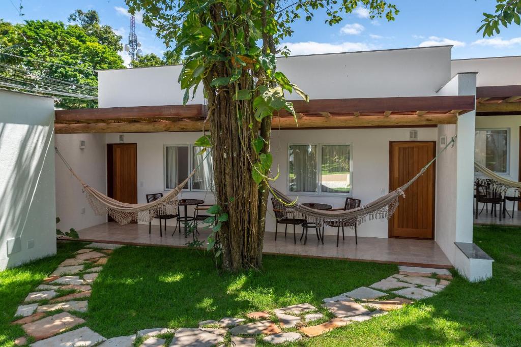 Edifício branco moderno com duas portas de madeira, redes penduradas em uma varanda e uma grande árvore na frente em um gramado, do Charm Bahia Suites. Representa hotéis em Arraial d’Ajuda.