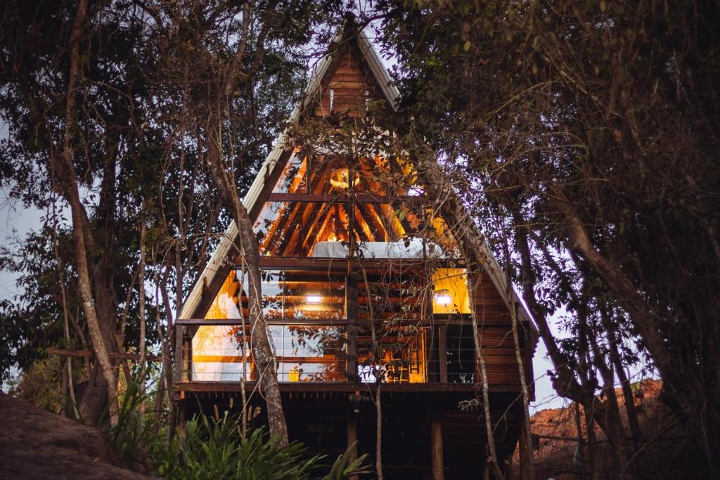 Cabana de madeira com janelas grandes, cercada por árvores, iluminada por luzes quentes à noite, em Entre Serras & Águas. Representa hotéis em Capitólio.