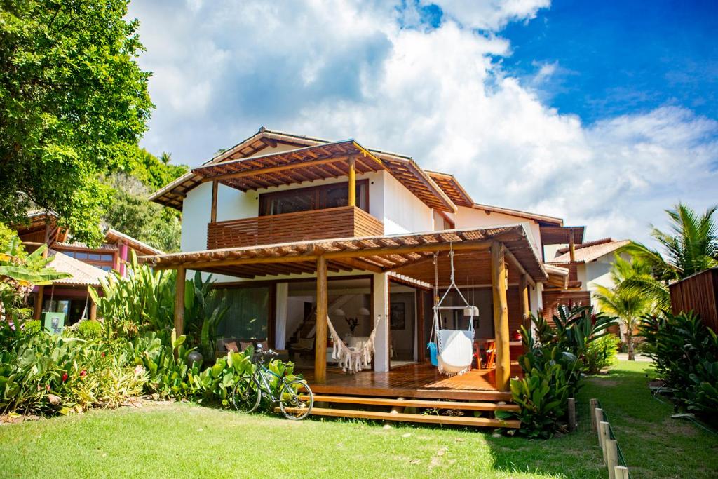Casa de dois andares com detalhes em madeira, varanda coberta, redes e paisagismo exuberante sob um céu parcialmente nublado, no Excelente casa, pé na areia, no melhor de Arraial!. Representa hotéis em Arraial d’Ajuda.