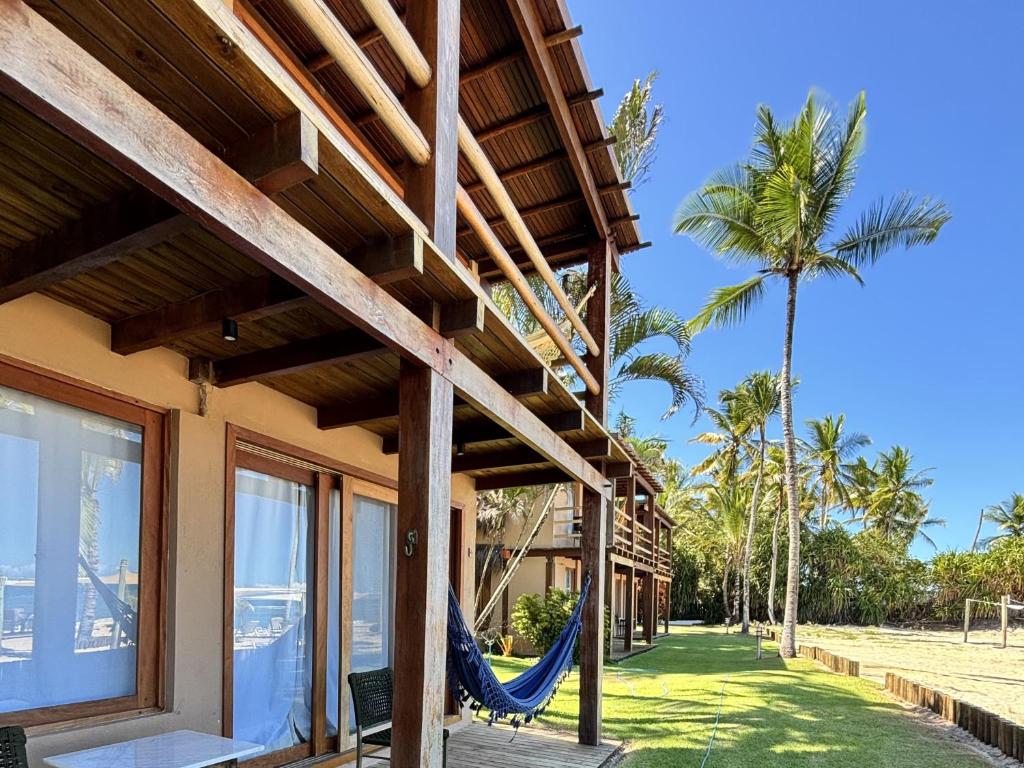 Edifício de madeira de dois andares à beira-mar com redes nos pátios, palmeiras e céu azul claro, do Hotel Pousada Pitinga. Representa hotéis em Arraial d’Ajuda.