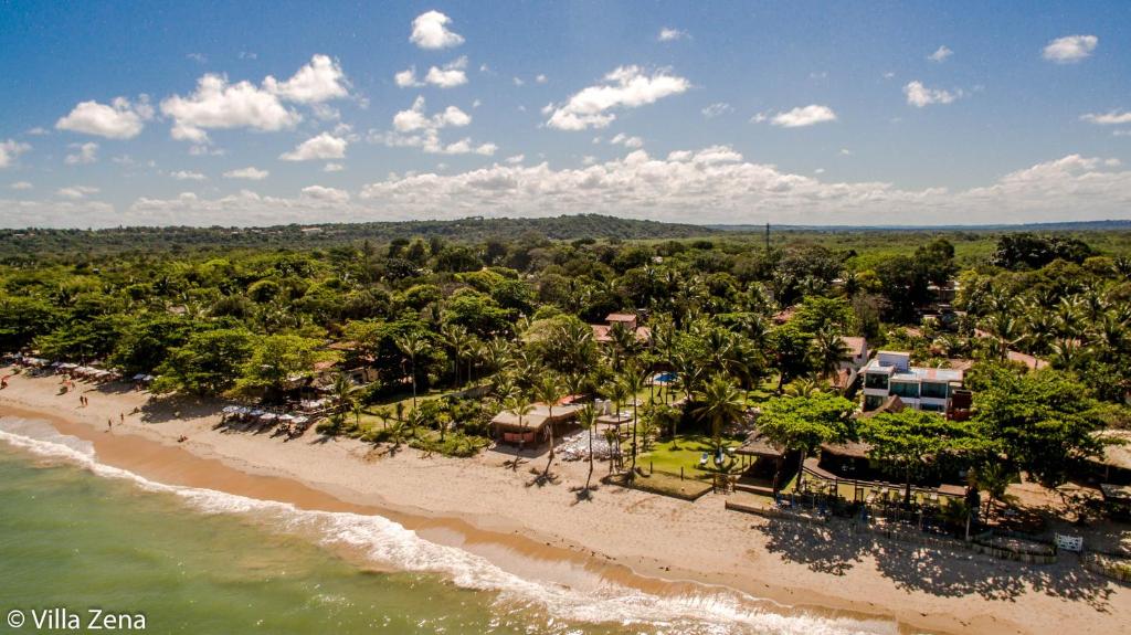 Vista aérea de uma praia de areia com ondas suaves e edifícios cercados por densas árvores verdes sob um céu parcialmente nublado, do Hotel Pousada Villa Zena. Representa hotéis em Arraial d’Ajuda.