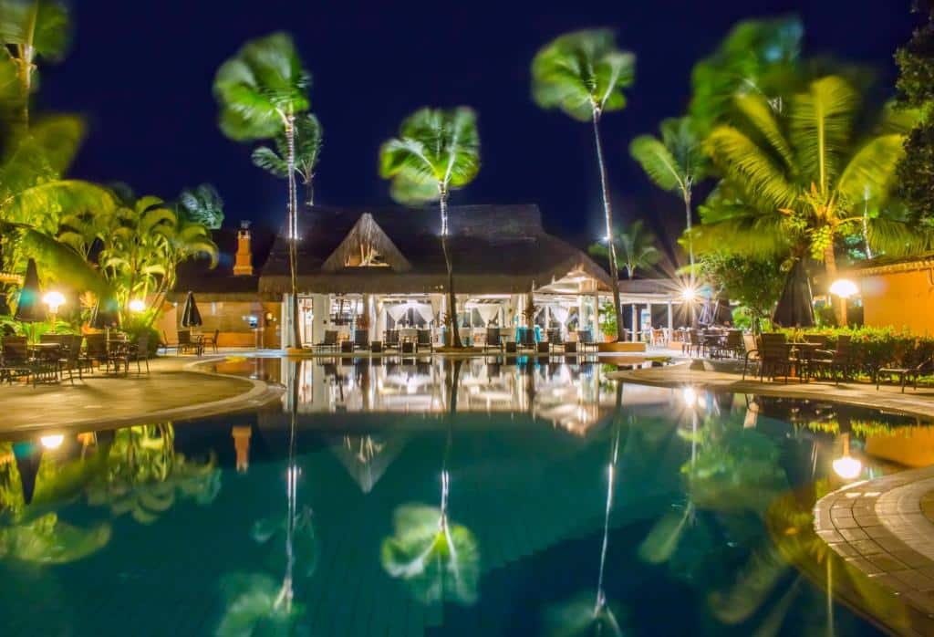 Um prédio de resort bem iluminado à noite é refletido em uma grande piscina cercada por palmeiras e assentos ao ar livre, do Mar Paraíso Hotel. Representa hotéis em Arraial d’Ajuda.