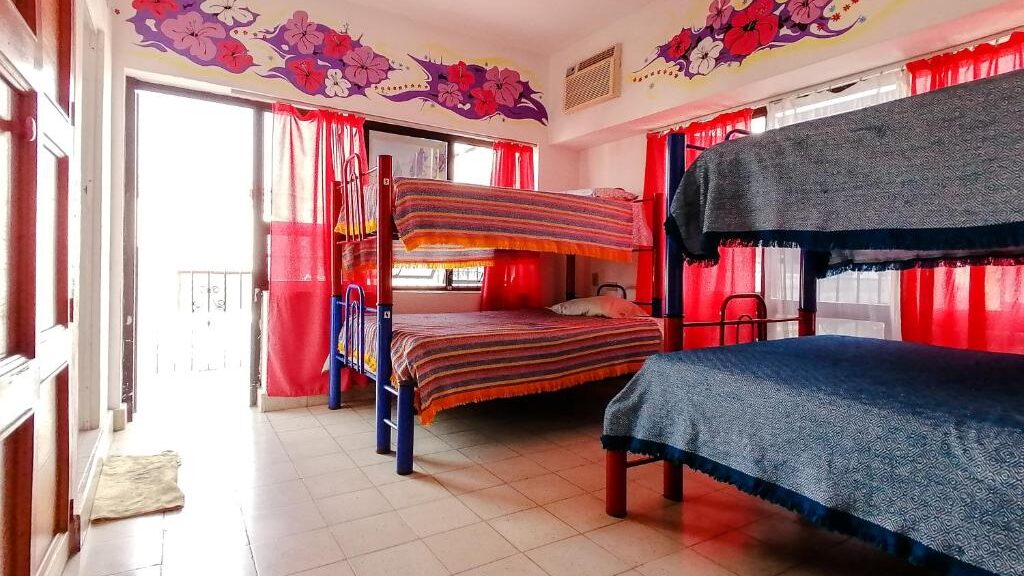 Um quarto do Hostel Vallarta bem iluminado com dois beliches, cortinas vermelhas, piso de ladrilho e mural floral acima das janelas e da porta. Essa é uma das dicas de onde ficar em Puerto Vallarta.