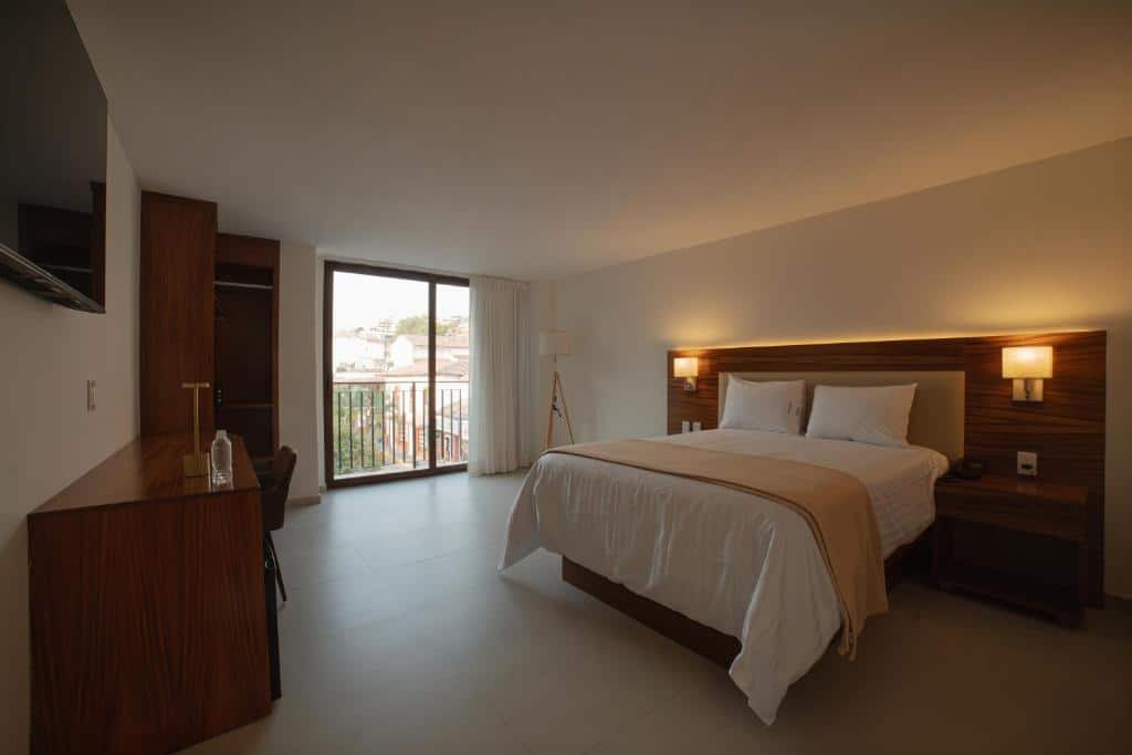 Um quarto do Hotel Casa Kavia Vallarta com uma cama de casal, móveis de madeira, escrivaninha, TV e uma porta de vidro deslizante que dá para uma varanda. Essa é uma das dicas de onde ficar em Puerto Vallarta.