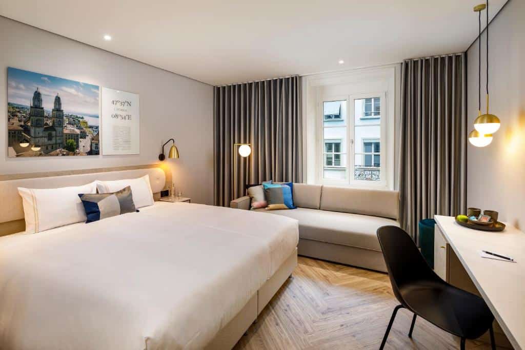 Hotel Felix, ilustra onde ficar em Zurique. Quarto de hotel moderno com uma cama de casal, um sofá perto da janela, uma escrivaninha com uma cadeira e arte na parede acima da cama.