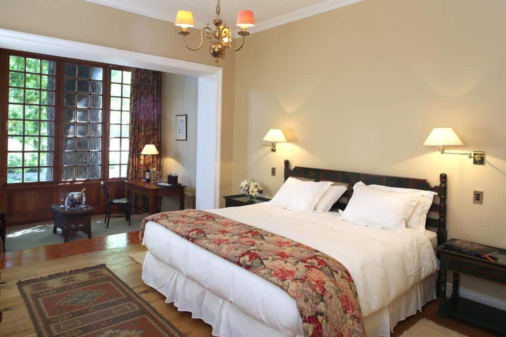Um quarto do Hotel Casa Real – Viña Santa Rita com uma cama grande, colcha floral, luminárias de parede, escrivaninha, janela com cortinas e piso de madeira.