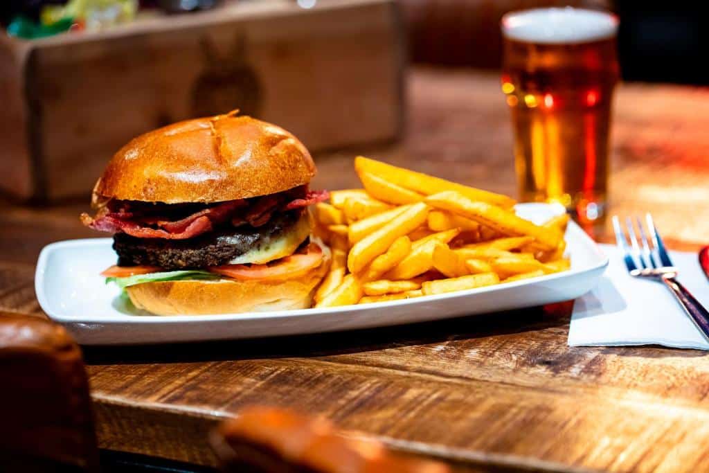 Kick Ass Grassmarket e Um cheeseburger com bacon, alface e tomate servido com batatas fritas em um prato branco ao lado de um copo de cerveja. Ilustra o post de onde ficar em Edimburgo.