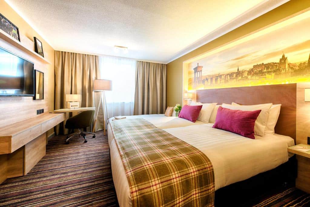 Quarto de hotel no Leonardo Hotel Edinburgh com uma cama de casal, cobertor xadrez, escrivaninha, TV, janela com cortinas e uma grande obra de arte de paisagem urbana acima da cama. Ilustra o post de onde ficar em Edimburgo.