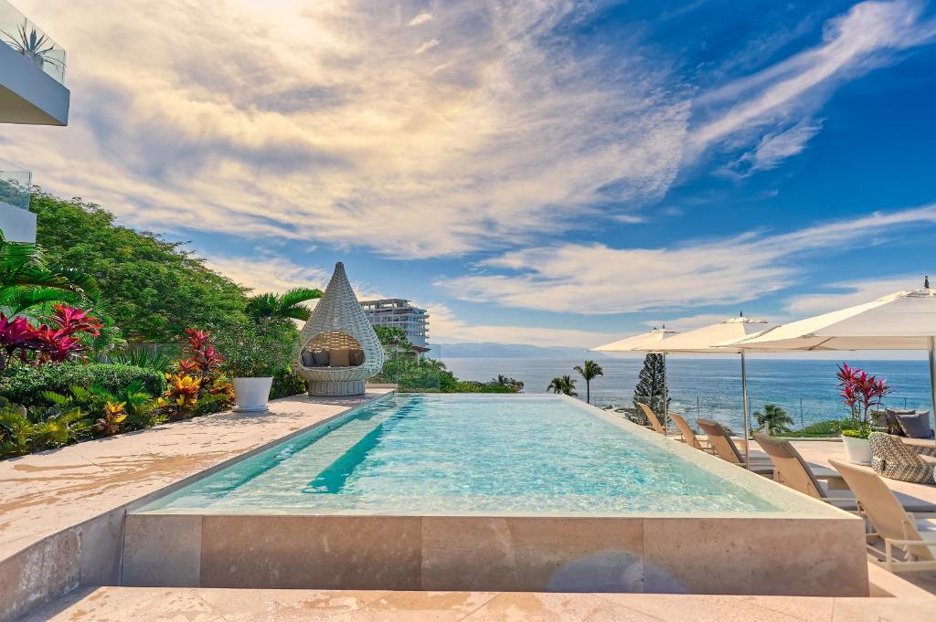 Piscina infinita das Maxwell Residences at Indah com espreguiçadeiras e guarda-sóis com vista para o oceano, cercada por plantas tropicais sob um céu parcialmente nublado.