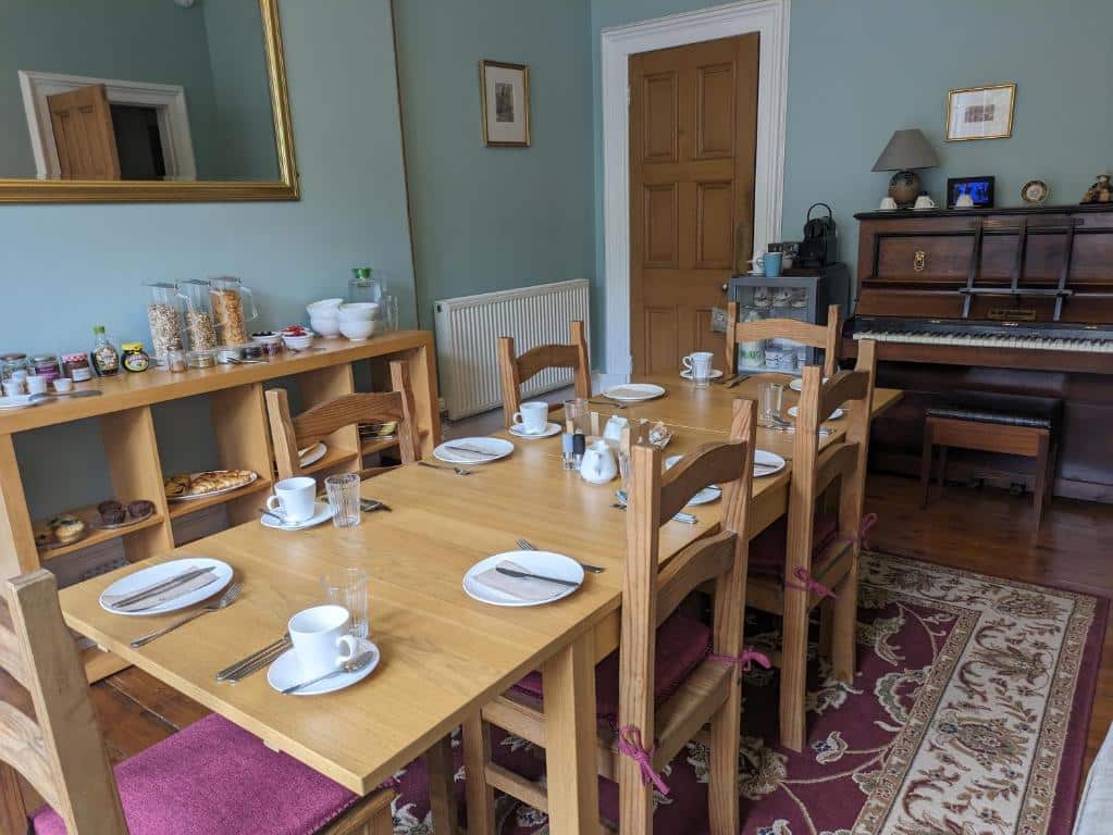 Uma sala de jantar no McCrae’s Bed and Breakfast com uma mesa de madeira posta, seis cadeiras, alimentos para o café da manhã em uma mesa lateral e um piano vertical no canto.