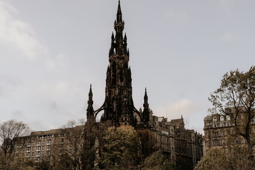 Monumento Scott gótico alto e escuro está entre as árvores, com edifícios históricos ao fundo sob um céu nublado.