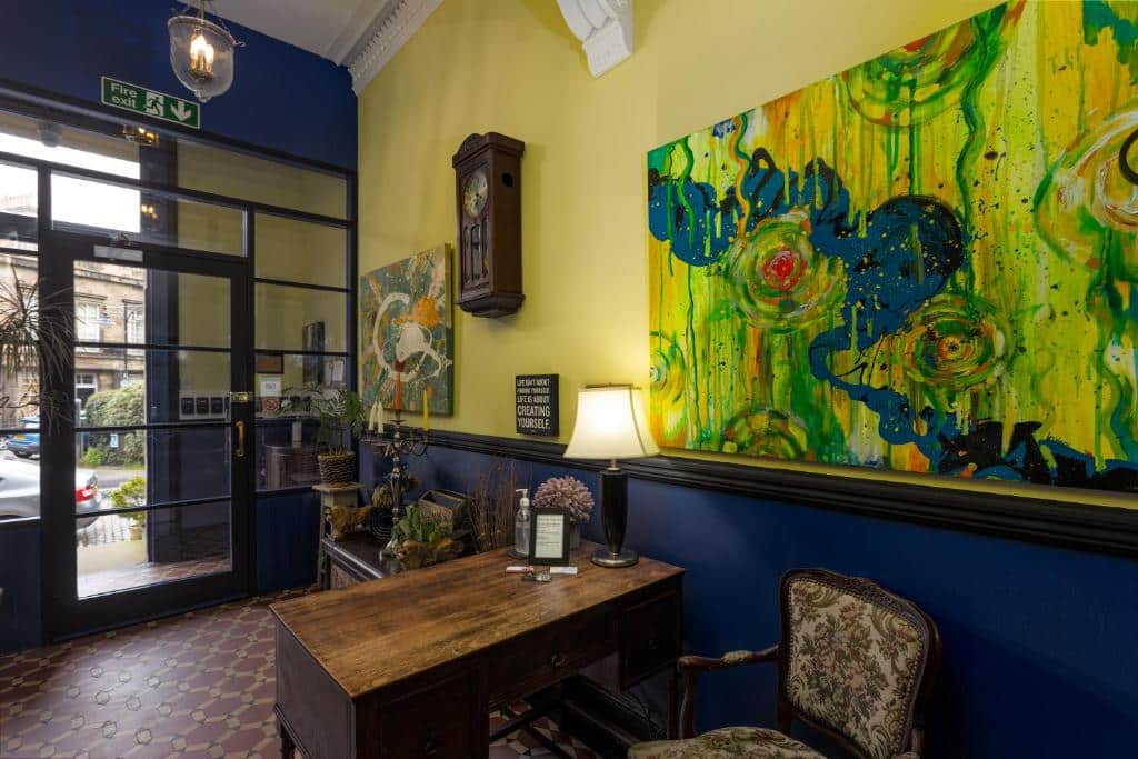 Uma pequena área de recepção no New Town Guest House com uma mesa de madeira, cadeira ornamentada, relógio de parede, luminária e pinturas abstratas coloridas nas paredes.