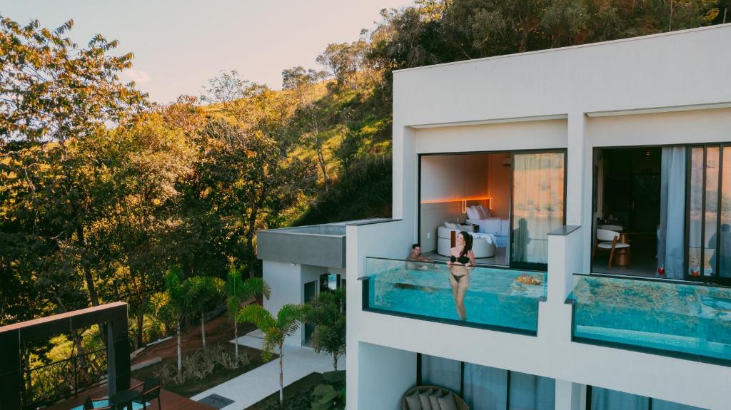 Uma pessoa em traje de banho está em uma piscina com paredes de vidro em uma varanda anexa a um moderno edifício branco cercado por árvores, no Collegare Hotel. Representa hotéis em Capitólio.